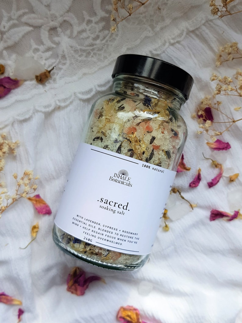 Puede incluir: Un frasco de vidrio lleno de una mezcla de sal de ba&ntilde;o blanca con flores y hierbas secas. La etiqueta dice "INHALE Botanicals .sacred. soaking salt 100% natural" y enumera los ingredientes como aceites esenciales de lavanda, cipr&eacute;s y romero. La etiqueta tambi&eacute;n indica que la mezcla est&aacute; dise&ntilde;ada para ayudar a restaurar la mente y recuperar el enfoque cuando te sientes abrumado. El frasco contiene 240 gramos de sal de ba&ntilde;o.