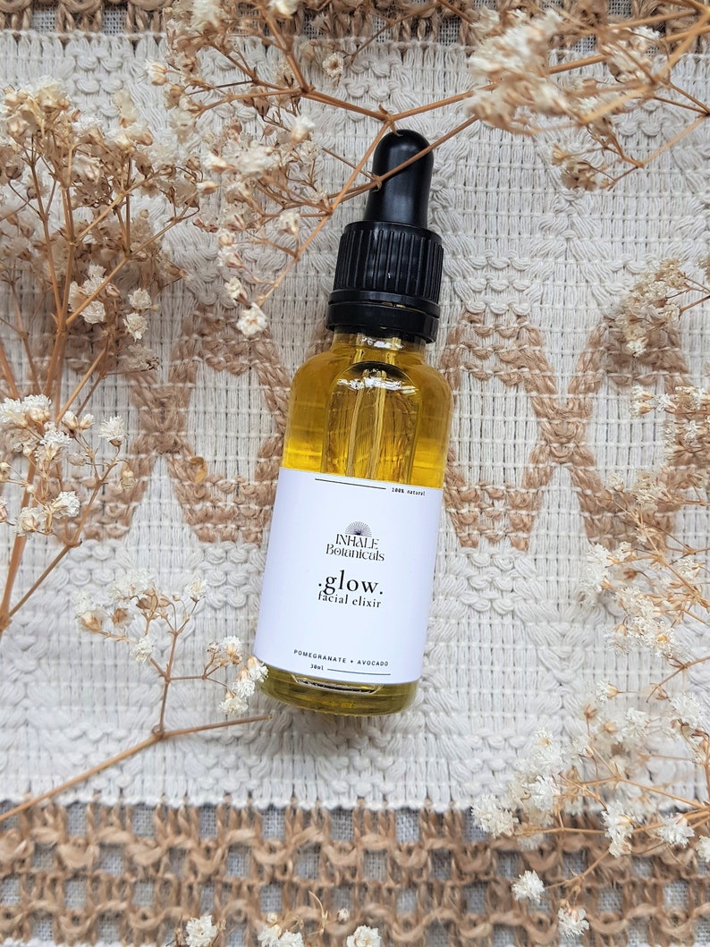 May include: A glass bottle of facial elixir with a black dropper top. The bottle is labeled "INHALE Botanicals .glow. facial elixir POMEGRANATE + AVOCADO 30ml".