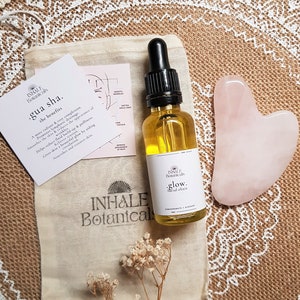 May include: A set of beauty tools for facial massage and skincare. The set includes a gua sha tool, a bottle of facial elixir, and a small pouch. The gua sha tool is made of rose quartz and is heart-shaped. The bottle of facial elixir is clear glass with a gold label that reads "INHALE Botanicals .glow. facial elixir". The pouch is made of natural fabric and has the brand name "INHALE Botanicals" printed on it.