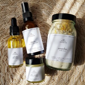 May include: Four glass bottles with labels containing natural beauty products. The products are labeled "glow. facial elixir", "nuit. sacred space mist", "oh rosa. treatment balm", and "soothe. milk bath".