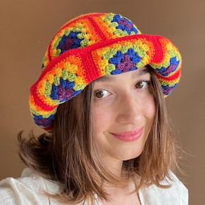 May include: A crochet bucket hat constructed from multi-colored granny squares featuring vibrant shades of red, orange, yellow, green, blue, and purple. The hat has a wide, flexible brim and is assembled with bright red yarn borders between squares.