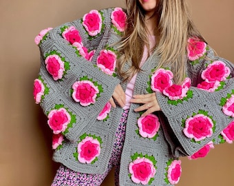May include: A gray, open-front crochet cardigan adorned with large, vibrant pink and green floral appliques. The cardigan features a square pattern and wide sleeves. The model is wearing floral print pants.