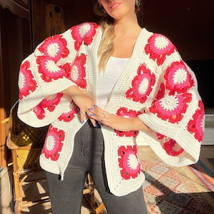 May include: A white crochet cardigan with pink and red flower accents. The cardigan has a loose fit and long sleeves.