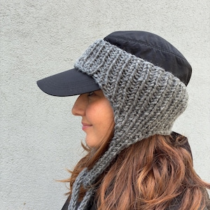Puede incluir: Una gorra de béisbol negra con una gruesa pieza de lana gris tejida a mano que cubre las orejas, los lados y la parte posterior. El componente tejido incluye largos cordones trenzados que cuelgan de los lados de la gorra.
