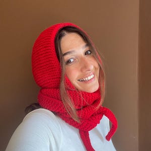 May include: A bright red knitted balaclava hood with an integrated scarf design. The accessory features a thick, textured ribbed knit pattern that covers the head and wraps around the neck, with two long knitted ties hanging down from the front.