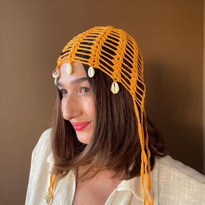 May include: A bright orange crochet headpiece featuring an open-knit pattern and dangling white cowrie shells attached along the front edge and sides. The accessory includes long, thin crocheted ties hanging down from the sides of the head.