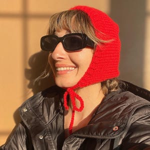 May include: A bright red crocheted bonnet with a textured knit pattern and thin chin ties. The bonnet is worn with black rectangular sunglasses and a black quilted jacket featuring silver-toned metal snap buttons along the front closure.