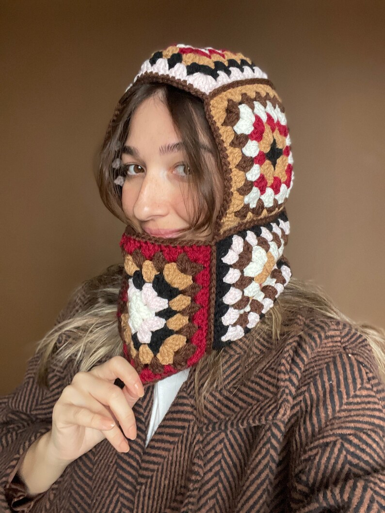 May include: A brown, black, white, and red crocheted hat with a hood and a neck warmer. The hat is made of granny squares.
