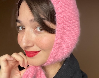 May include: A bright pink knitted bonnet with a textured ribbed border along the face opening. The headpiece features a soft, fuzzy yarn construction and includes two knitted ties that hang down from the chin area to secure the bonnet in place.