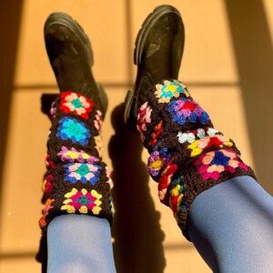 May include: A pair of leg warmers crafted from black yarn featuring a multi-colored granny square pattern. The squares incorporate vibrant shades of blue, pink, yellow, and red. These crochet leg warmers are worn over blue tights and black boots.