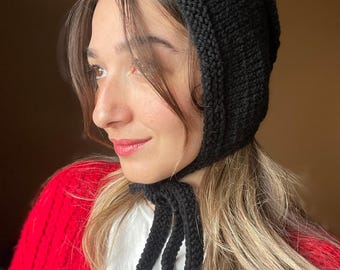 May include: A solid black knitted bonnet featuring a textured garter stitch border along the face opening and two long knitted ties that fasten under the chin. The headwear is crafted from a thick yarn with a visible knit pattern throughout the fabric.