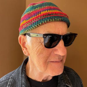 Peut inclure: Un bonnet en crochet multicolore avec des rayures horizontales dans des tons de rouge, jaune, vert et bleu. Le bonnet présente une texture en maille serrée et une forme arrondie. L'accessoire est associé à des lunettes de soleil noires.