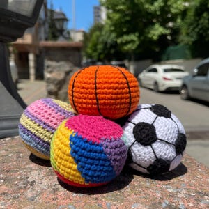 May include: A collection of five colorful, crocheted balls. The balls include a basketball, a soccer ball, and three other balls with various color patterns. The balls are sitting on a weathered, reddish-brown surface.
