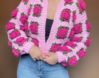 May include: A light pink crochet cardigan featuring three-dimensional dark pink rose appliqués with green leaves. The garment is constructed from square panels with a ribbed texture along the front opening and cuffs, worn over a black top and blue jeans.