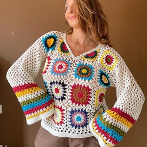 Può includere: Un top all'uncinetto a maniche lunghe con base bianca, scollo a V e un vivace motivo a piastrelle granny. Le maniche presentano strisce orizzontali gialle, verdi, blu e rosa, mentre il corpo mostra motivi floreali multicolori.