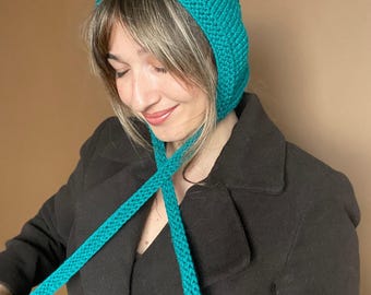 May include: A teal knitted bonnet featuring a textured ribbed pattern and long, matching knitted ties that extend downwards. The headwear is constructed from a thick yarn, providing a structured shape that covers the top and sides of the head.