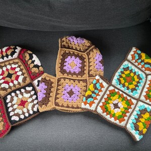 May include: Three crocheted granny squares in different colors and patterns. The squares are brown, purple, and blue with white and yellow accents. The squares are joined together to form a larger piece.