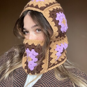 May include: A brown and purple crocheted hat with a flap that covers the face. The hat is made of granny squares with a purple flower design.