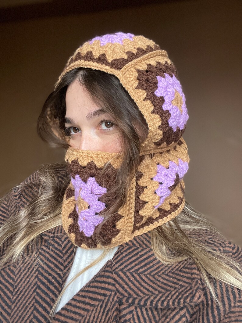 May include: A brown and purple crocheted hat with a flap that covers the face. The hat is made of granny squares with a purple flower design.