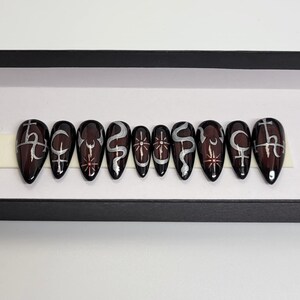 May include: Set of ten black and red coffin-shaped press-on nails with silver designs. The designs include a crescent moon, a snake, a cross, and a star.
