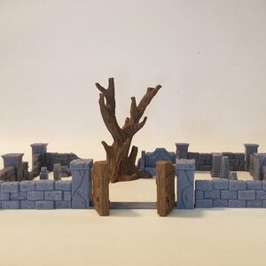 Może przedstawiać: Miniaturowa kamienna ściana z drewnianą bramą i martwym drzewem w środku. Ściana jest wykonana z szarego kamienia i ma postarzały wygląd. Brama jest wykonana z brązowego drewna i ma prosty wzór. Drzewo jest wykonane z brązowego plastiku i ma powykręcany, sękowy pień. Zestaw idealnie nadaje się do gier planszowych lub miniaturowych dioram.