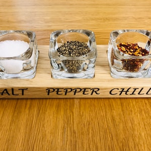 May include: Wooden spice rack with three glass jars labelled "SALT", "PEPPER", and "CHILLI". The jars are filled with white salt, black peppercorns, and red chilli flakes.