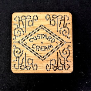 May include: A wooden coaster featuring a black design of a diamond shape with the words "Custard Cream" inside. The diamond is surrounded by four decorative swirls.
