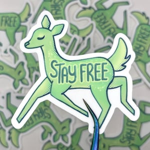 Puede incluir: Una pegatina verde con un ciervo estilizado con la frase "STAY FREE" en su interior. El ciervo está en posición de carrera, con un efecto degradado y un borde blanco. Otras pegatinas con el mismo diseño están en el fondo.