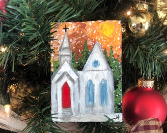 Puede incluir: Un adorno navideño pintado a mano que representa una iglesia blanca con una puerta roja y ventanas azules sobre un fondo de puesta de sol naranja con nieve.