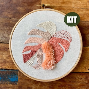 Può includere: Un kit di ricamo con un design a forma di foglia di monstera. La foglia è cucita con varie trame e tonalità di pesca, crema e bordeaux. Il disegno è inserito in un telaio da ricamo in legno, con un cerchio verde che dice "KIT".