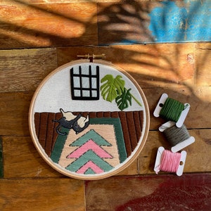May include: Embroidered hoop art featuring a black cat on a pink and green rug with a window and a potted plant in the background. The hoop is surrounded by spools of embroidery floss in pink, green, and grey.