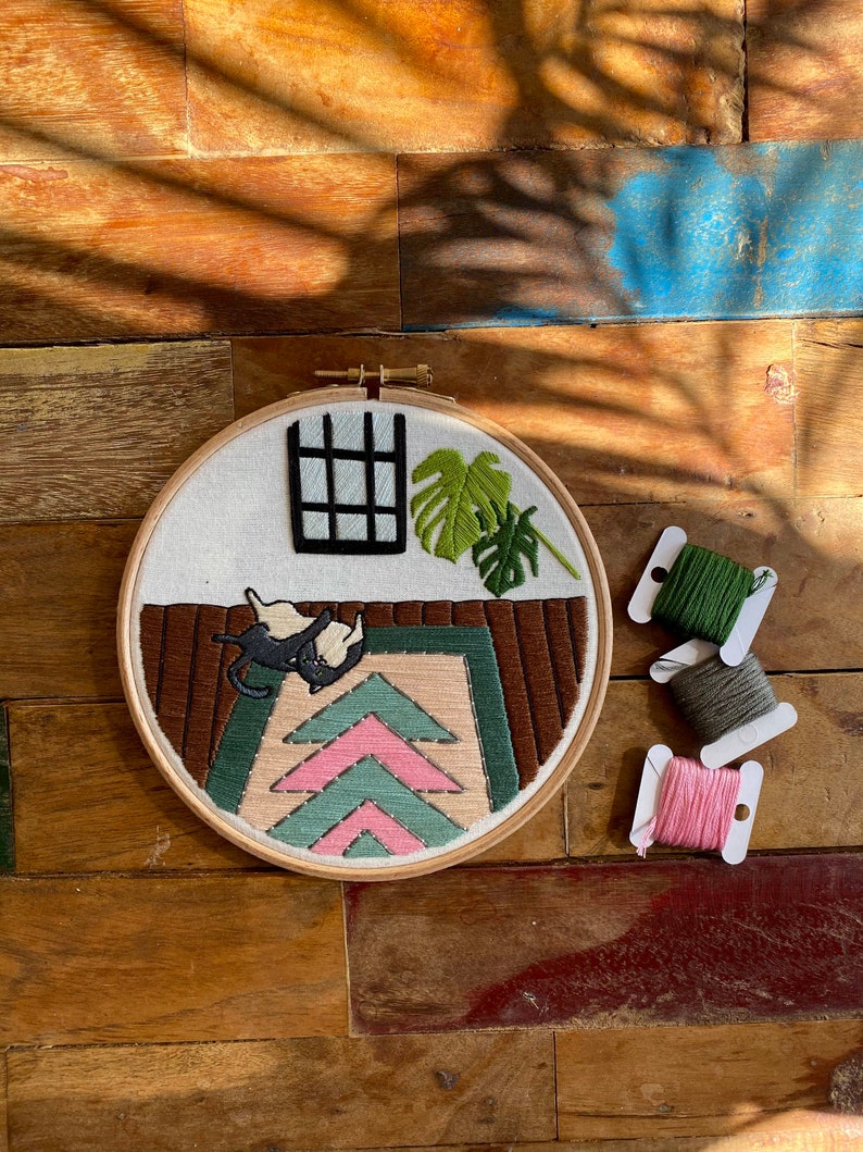 May include: Embroidered hoop art featuring a black cat on a pink and green rug with a window and a potted plant in the background. The hoop is surrounded by spools of embroidery floss in pink, green, and grey.