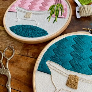 May include: Two embroidery hoops with white fabric and colourful embroidery designs. The top hoop features a pink and white geometric pattern, a white bathtub with a green plant hanging over it, and a teal blue circle. The bottom hoop features a teal blue geometric pattern, a white bathtub with a gold bar of soap, and a pink and white geometric pattern.