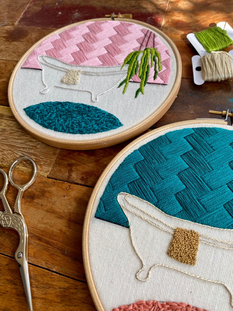 May include: Two embroidery hoops with white fabric and colourful embroidery designs. The top hoop features a pink and white geometric pattern, a white bathtub with a green plant hanging over it, and a teal blue circle. The bottom hoop features a teal blue geometric pattern, a white bathtub with a gold bar of soap, and a pink and white geometric pattern.