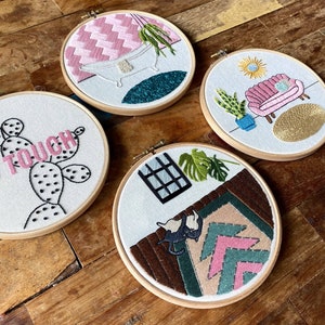 May include: Four embroidery hoops featuring different designs. The first hoop shows a pink and white checked background with a white bathtub and a green plant. The second hoop has a white background with a pink sofa, a green plant, and a yellow sun. The third hoop has a white background with a cactus and the word "TOUCH". The fourth hoop has a brown background with a pink and green geometric pattern and a black cat.