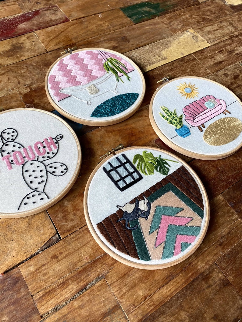 May include: Four embroidery hoops featuring different designs. The first hoop shows a pink and white checked background with a white bathtub and a green plant. The second hoop has a white background with a pink sofa, a green plant, and a yellow sun. The third hoop has a white background with a cactus and the word "TOUCH". The fourth hoop has a brown background with a pink and green geometric pattern and a black cat.