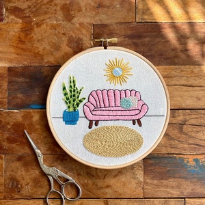May include: Embroidered hoop art featuring a pink sofa with a blue cushion, a green plant in a blue pot, a yellow sun, and a beige rug.