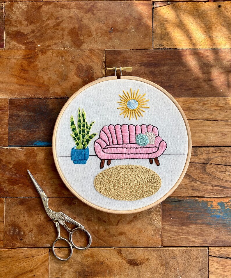 May include: Embroidered hoop art featuring a pink sofa with a blue cushion, a green plant in a blue pot, a yellow sun, and a beige rug.