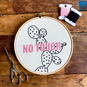 May include: Embroidered hoop art featuring a cactus design and the text "NO TOUCH" in pink thread. The hoop is on a wooden surface with black and pink embroidery floss.