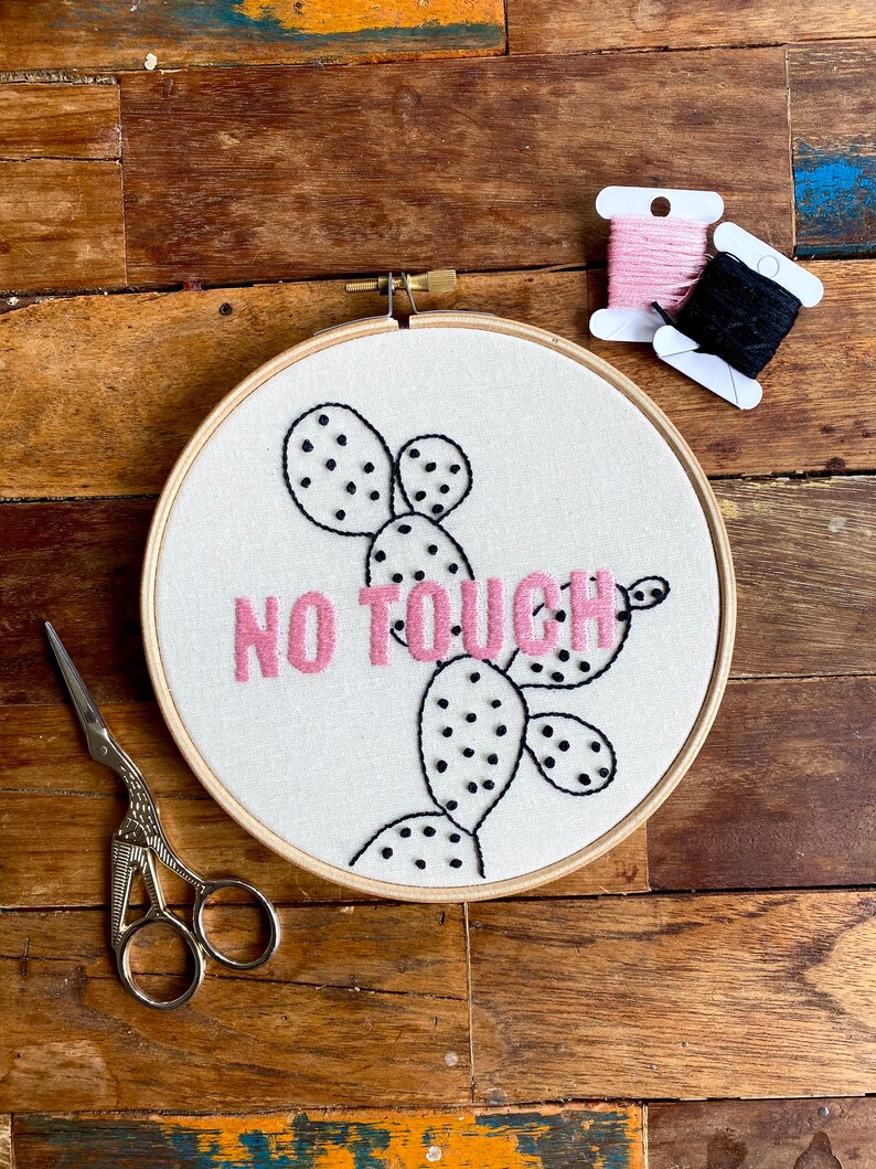 May include: Embroidered hoop art featuring a cactus design and the text "NO TOUCH" in pink thread. The hoop is on a wooden surface with black and pink embroidery floss.