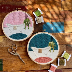 May include: Two embroidery hoops featuring a white bathtub, green plant, and pink and blue backgrounds. The hoops are on a wooden surface with spools of embroidery floss.