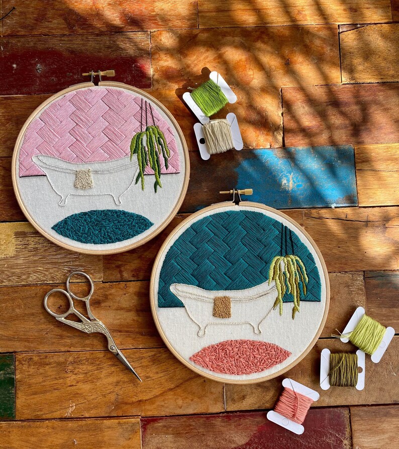 May include: Two embroidery hoops featuring a white bathtub, green plant, and pink and blue backgrounds. The hoops are on a wooden surface with spools of embroidery floss.
