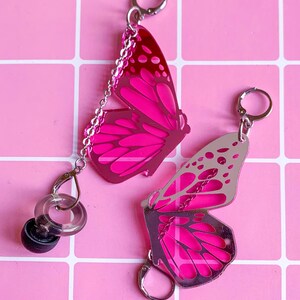 Op de afbeelding: Twee roze vlindervormige sleutelhangers met zilverkleurige hardware. Eén sleutelhanger heeft een volledig vlinderontwerp, de andere een halve vlinder. De sleutelhangers liggen op een roze rasterachtergrond.