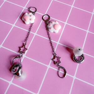Könnte beinhalten: Ohrringe mit weißen und rosa Katzenpfoten-Anhängern, silbernen Sternen und silbernen Ohrhaken. Ein einzelner silberner Ohrring mit einem weißen Ohrhörer ist ebenfalls vorhanden. Die Ohrringe werden auf einem rosa Gitterhintergrund präsentiert.