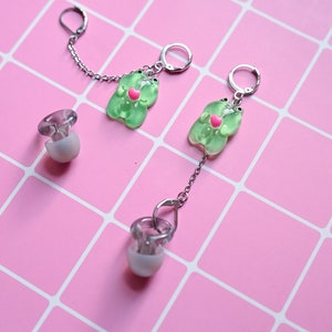 Könnte beinhalten: Ein Paar grüne Gummibärchen-Ohrringe mit einem rosa Herzen auf jedem Bären. Die Ohrringe sind durch eine silberne Kette verbunden.