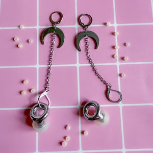 Könnte beinhalten: Silberfarbene Ohrringe mit Mond-Design, verbunden durch eine Kette mit einem kreisförmigen Ohrplug. Die Ohrringe sind auf einem rosa karierten Hintergrund platziert, mit kleinen, cremefarbenen Perlen verstreut.