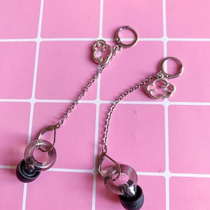 Puede incluir: Un par de pendientes plateados con un dije en forma de nube y un acento circular transparente. Los pendientes cuelgan de una cadena y presentan un componente negro similar a un auricular. Fondo rosa con patrón de cuadrícula.