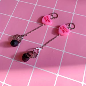 Puede incluir: Pendientes con colgantes rosas en forma de corazón con la inscripción "Sweet Heart" en blanco. Cuentan con cadenas plateadas y detalles de cuentas oscuras y transparentes. Fondo rosa con patrón de cuadrícula.