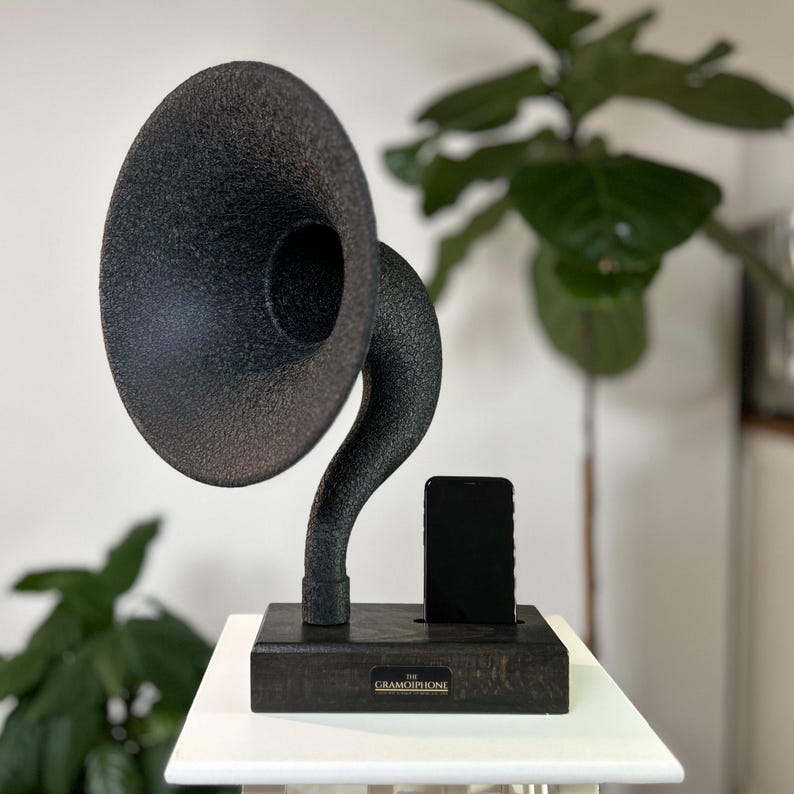 May include: A black vintage-style gramophone speaker with a horn shape and a wooden base. The speaker has a slot for a smartphone, and the base has the text "The Gramophone" on it.