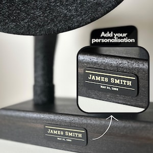 May include: Black wooden stand with a round black top. The stand has a personalised nameplate with the text "JAMES SMITH MAY 31, 1963".